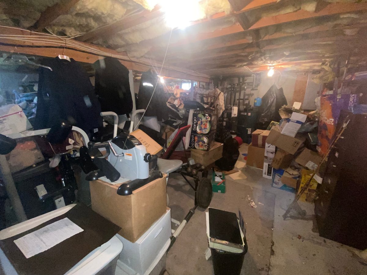 Wide view of a basement crowded with boxes, exercise equipment, and clothing before a cleanout