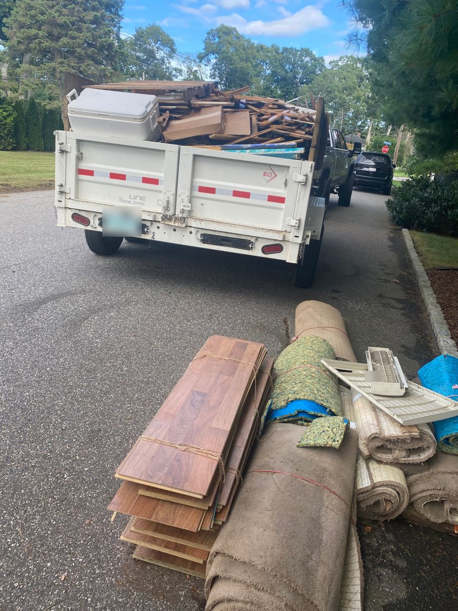 Dump trailer loaded with wood debris next to rolled carpet and flooring at the curb