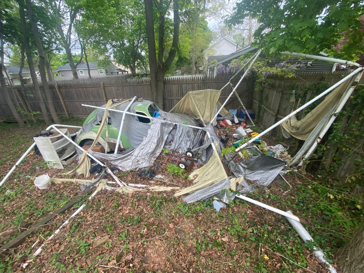 Collapsed canopy frame and tarps over clutter in an overgrown backyard